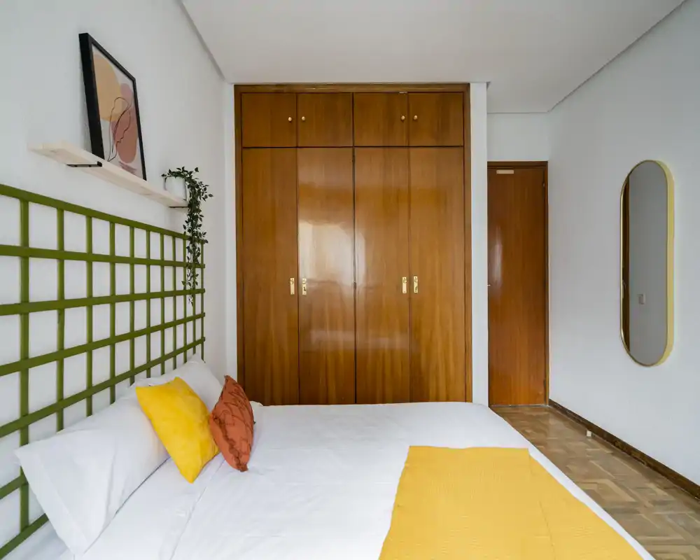 Chambre lumineuse et stylée avec lit blanc rehaussé d'accents jaunes et orange, tête de lit en treillis vert et placard mural en bois — lignes nettes et tons chauds parfaits pour une annonce.