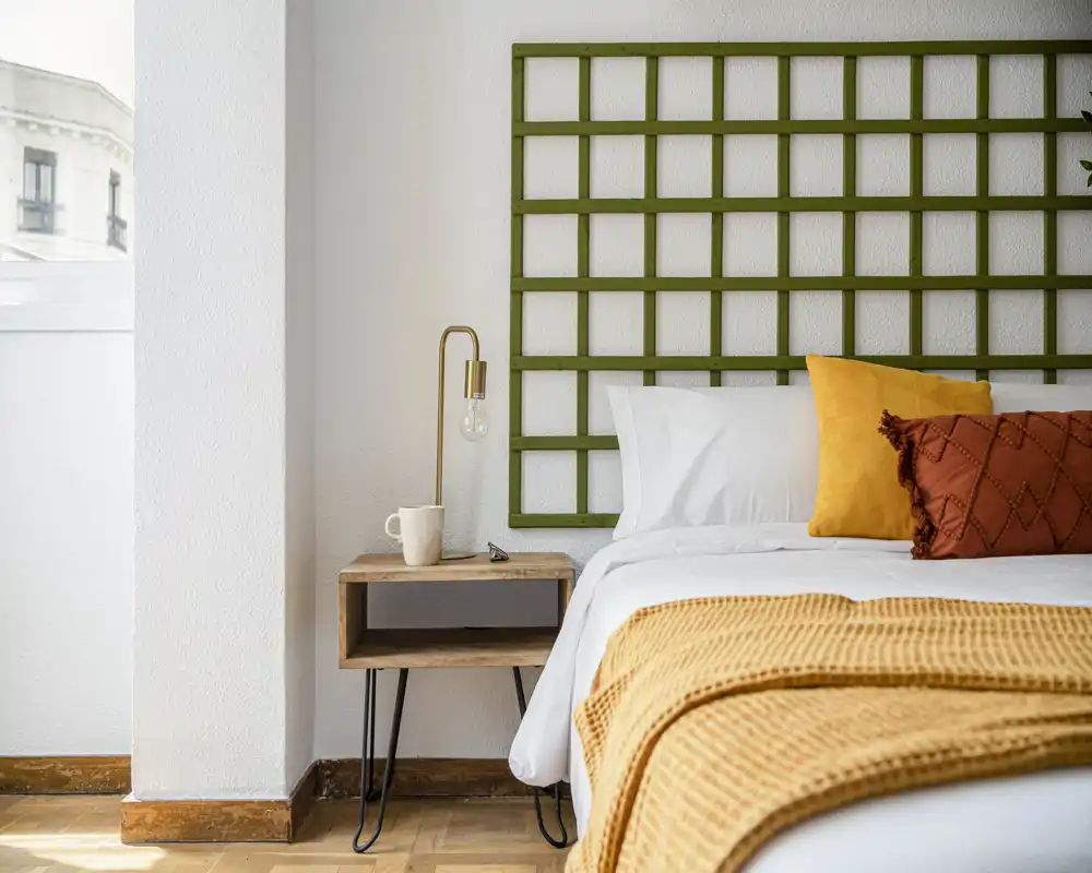 Coin chambre lumineux et soigné avec lit bien fait, plaid moutarde chaleureux et coussins décoratifs, table de chevet et lampe moderne près de la fenêtre.
