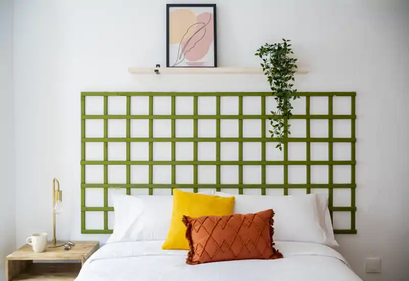 Chambre lumineuse et soignée avec un lit complet, des coussins décoratifs aux tons chauds et une tête de lit en treillis vert ; linge blanc propre et petite table en bois pour une ambiance moderne et accueillante.