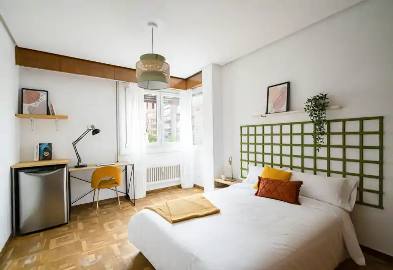Chambre lumineuse et soignée avec lit double, tête de lit décorative, table de chevet et un petit bureau près de la fenêtre — idéale pour se reposer et travailler à distance.
