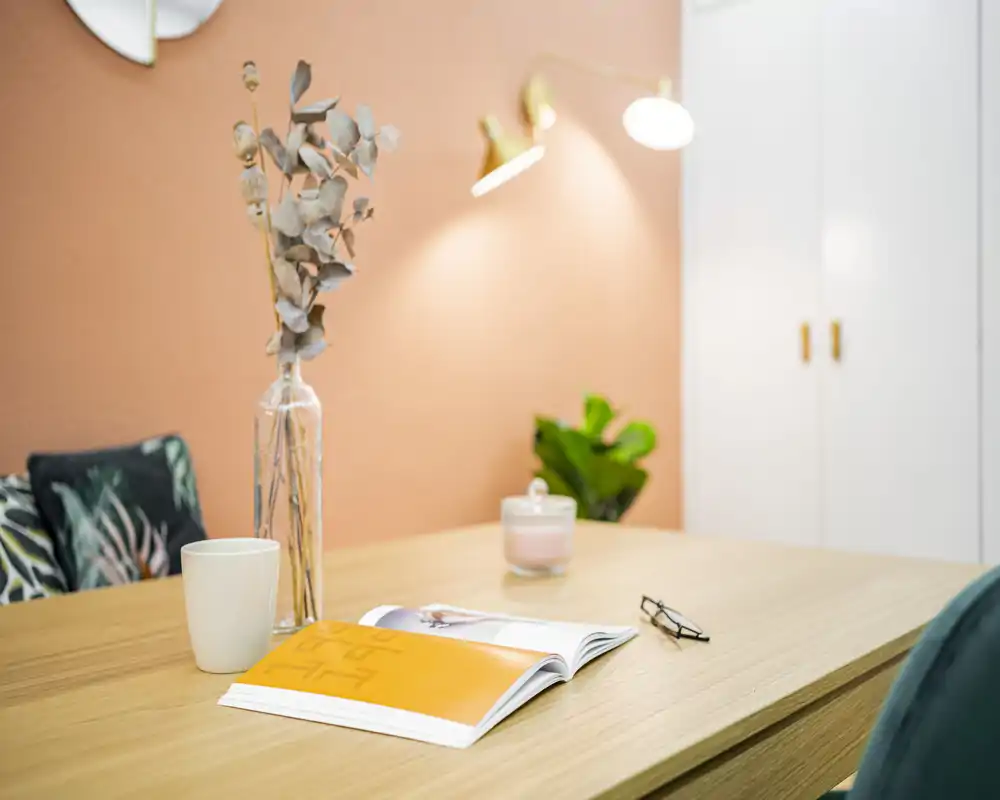 A stylish, warmly lit living area with a wooden table set against a peach wall, decorative cushions, a plant and soft lamp lighting — ideal for relaxed reading or coffee.