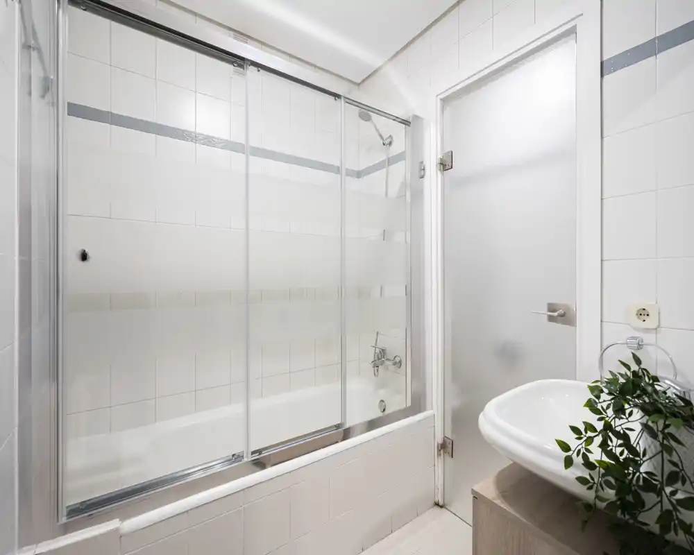 Clean, bright bathroom with a bathtub and sliding glass shower doors, white tiled walls and a modern sink with a small plant for a fresh touch.