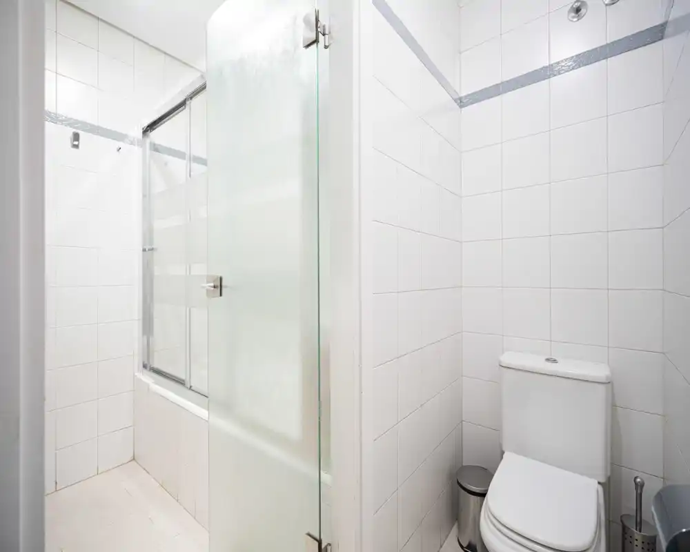 Clean, bright bathroom with white tiled walls, a toilet, and a shower/bathtub area with frosted glass door.