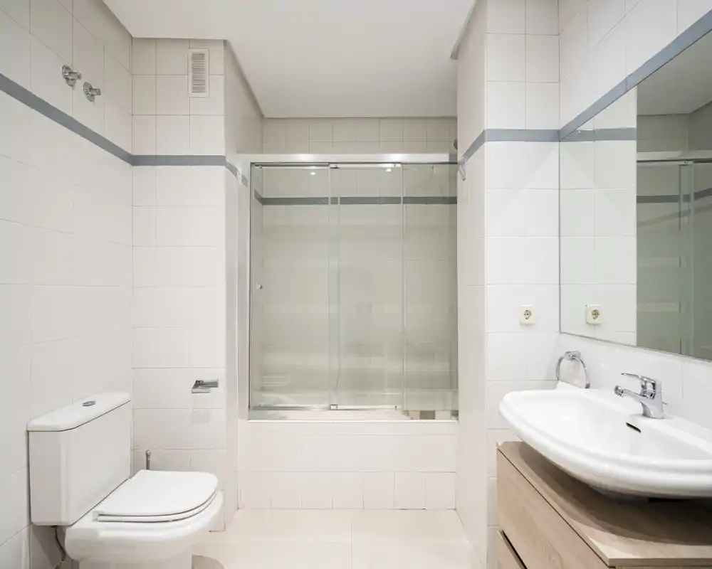 Clean, modern bathroom with a bathtub and sliding glass shower doors, wall-mounted mirror, sink with vanity, and a toilet. Bright neutral tiles and simple fixtures create a fresh, well-maintained space.