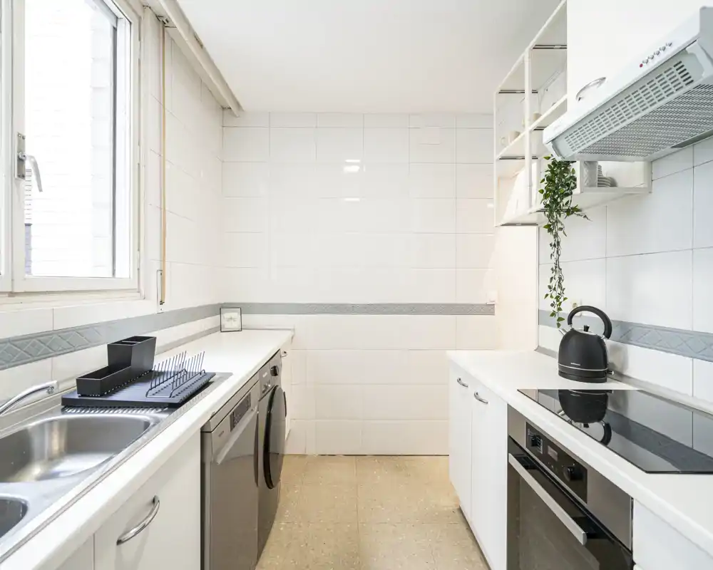 Bright, modern galley kitchen with white cabinetry, induction hob, built-in oven and dishwasher. Natural light from the window and minimalist accessories create a clean, functional space.