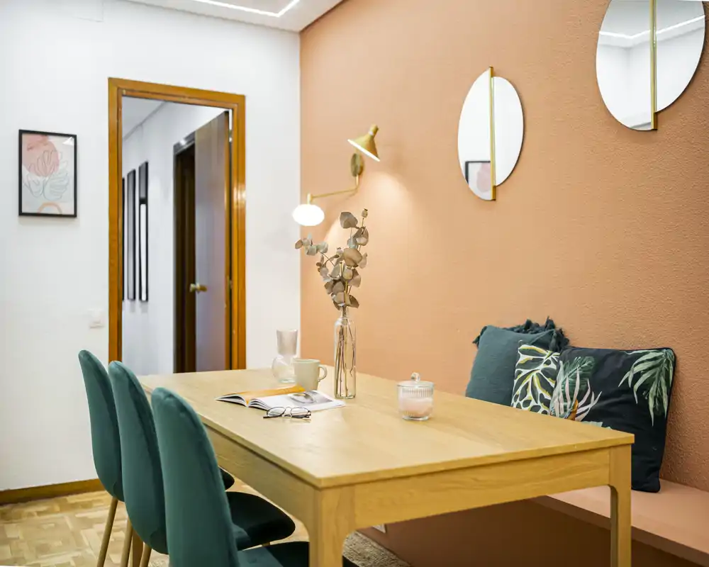 Stylish dining nook with a wooden table, green upholstered chairs and a cosy bench against a warm terracotta wall; decorative cushions, a vase and soft lighting create an inviting atmosphere for meals.