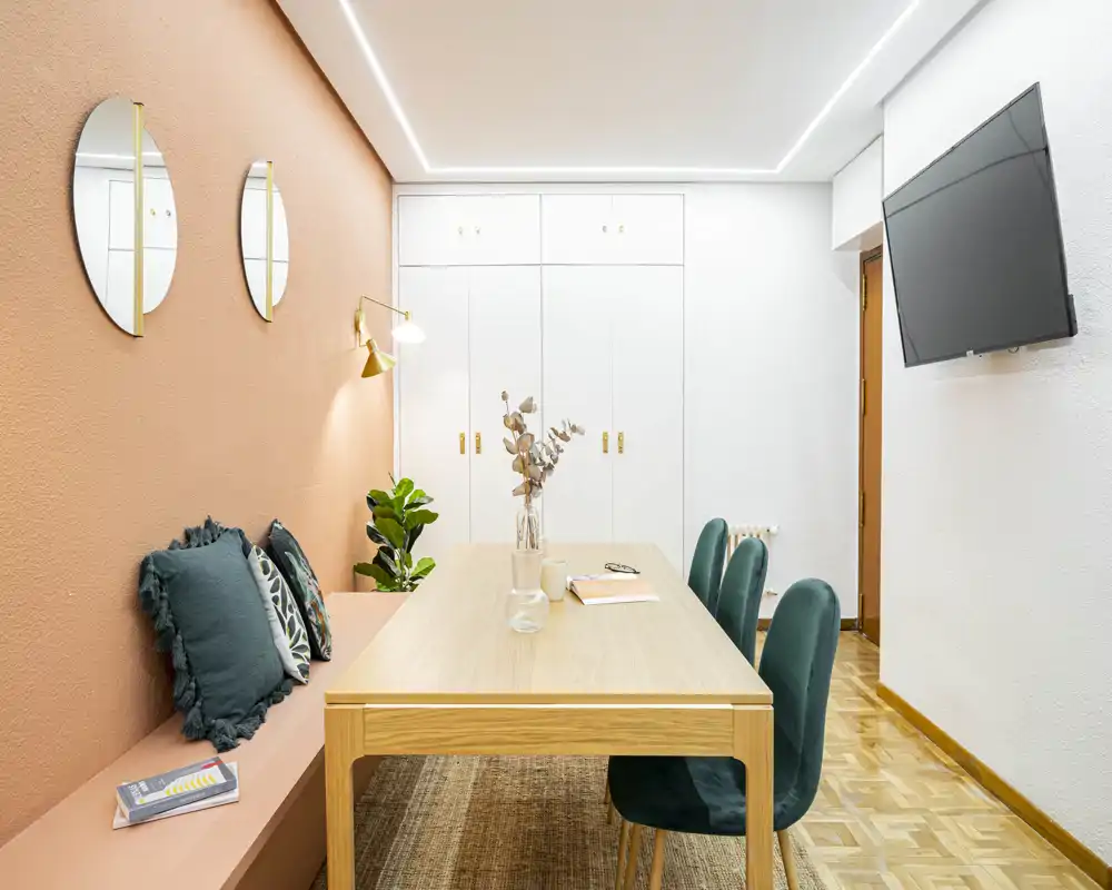 Stylish dining area with a light wood table, upholstered green chairs and a built-in bench against a warm terracotta wall. Clean contemporary styling, decorative cushions and plants create a welcoming spot for meals or work.