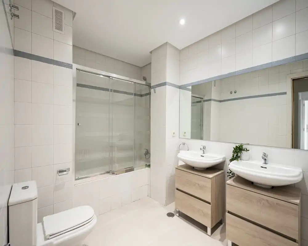 Bright, modern bathroom with a bathtub and glass shower screen, dual sink vanities with wooden drawers, large mirror and white tiled walls.