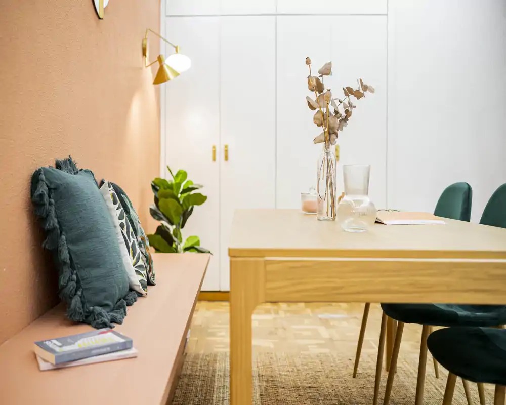 Bright, styled dining area with a wooden table, green velvet chairs and decorative cushions on a bench; warm colors and plants create a welcoming atmosphere.