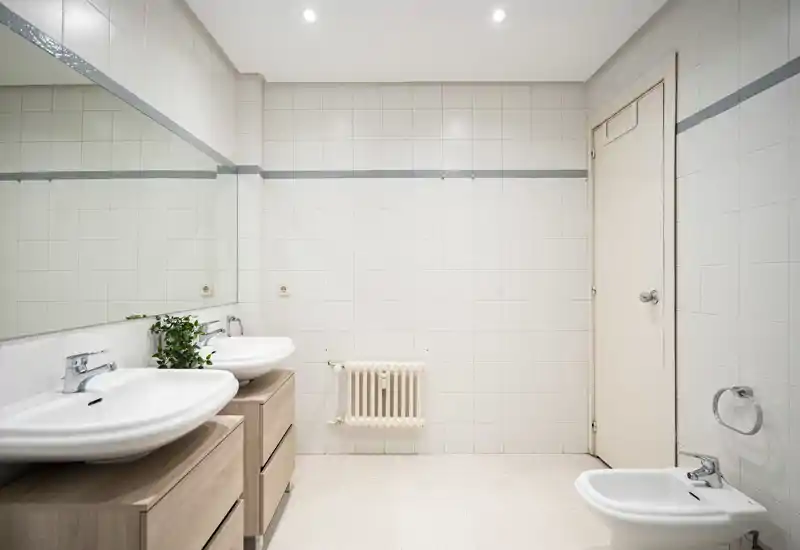 Salle de bains lumineuse et moderne avec doubles vasques sur meubles en bois, grand miroir, bidet et murs carrelés propres — idéale pour une routine matinale confortable.