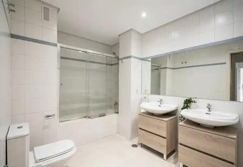 Salle de bains moderne et lumineuse avec baignoire et pare-douche en verre, deux vasques sur meubles en bois, grand miroir et carrelage blanc net.
