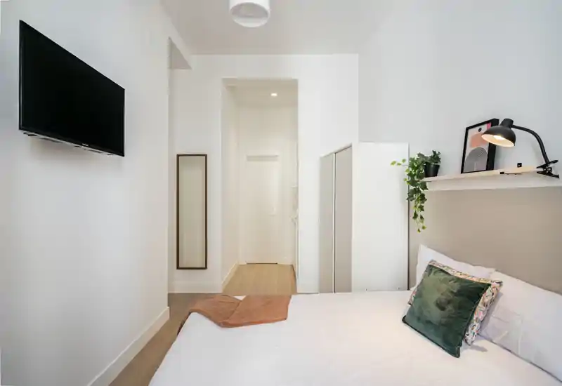 Dormitorio moderno y luminoso con cama doble bien hecha, cojines decorativos y lámpara de pared; armario y televisión visibles, decoración minimalista.