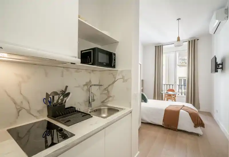 Camera luminosa e moderna in stile monolocale con angolo cottura compatto. Il letto è ben rifatto vicino a una grande finestra che illumina l’ambiente; un piccolo tavolo e una TV a parete completano la composizione accogliente.