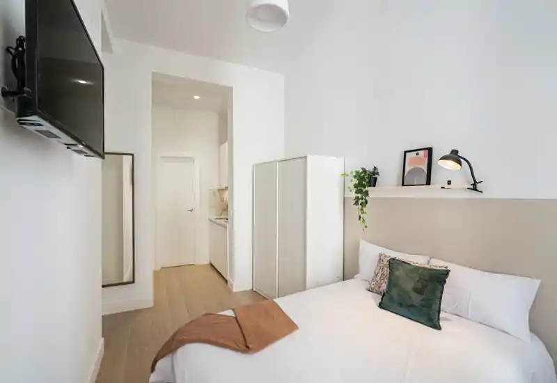 Dormitorio pequeño y luminoso con cama doble vestida con ropa blanca, cojines decorativos y una manta. La habitación incluye un armario, una repisa con planta y lámpara, televisión mural y un espejo de cuerpo entero junto a la entrada.