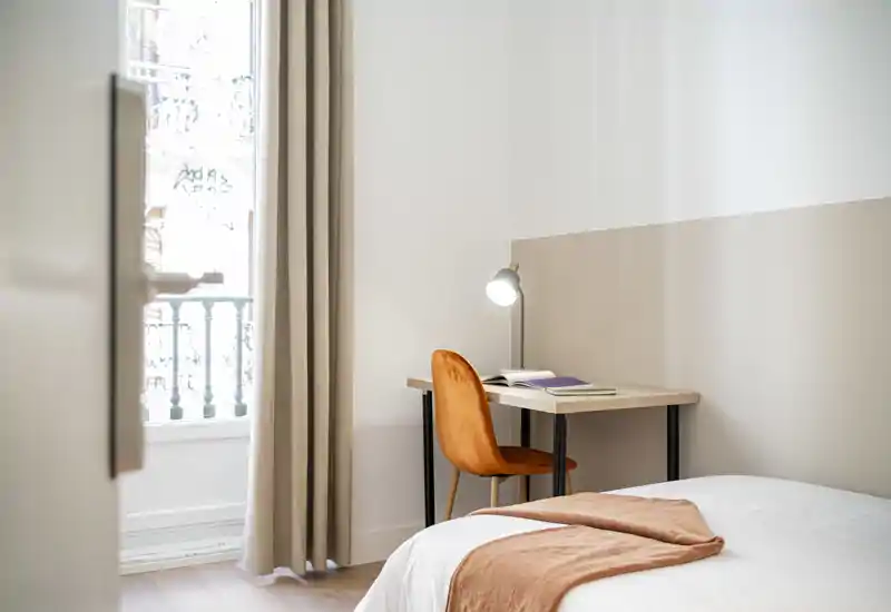 Camera da letto luminosa e moderna con letto ordinato, una coperta calda e una scrivania vicino alla finestra — ideale per lavorare e rilassarsi con luce naturale.