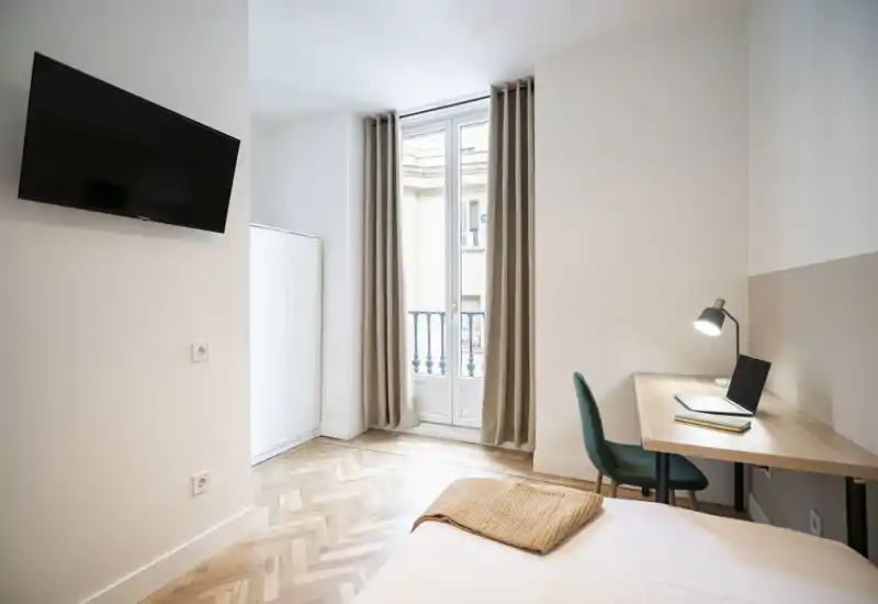 Camera da letto luminosa e moderna con letto ben rifatto, scrivania con lampada e sedia, pavimento in parquet e finestra alta con tende che garantisce luce naturale.