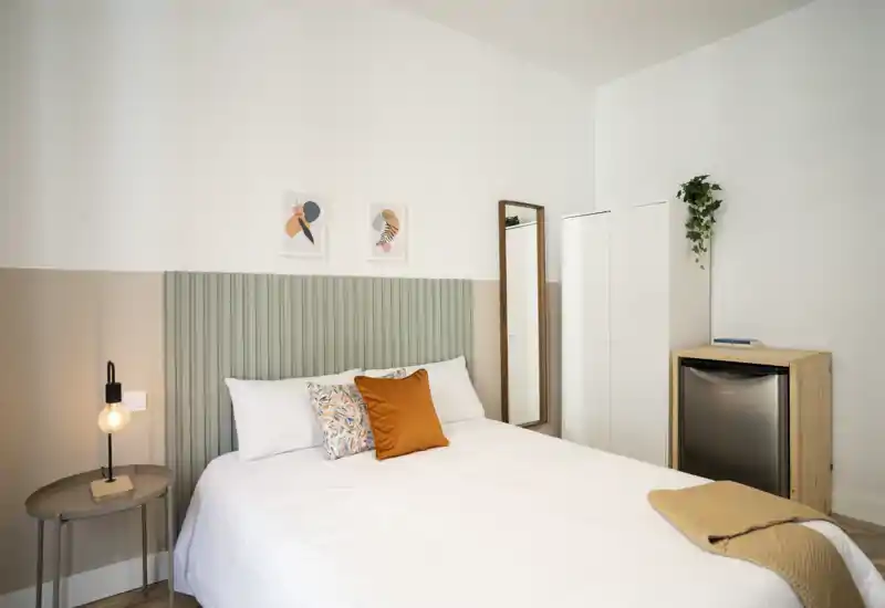Dormitorio luminoso y con estilo, con una cama bien hecha, cojines decorativos y una lámpara de mesita. La habitación dispone de un espejo de cuerpo entero, un armario compacto y un pequeño frigorífico.