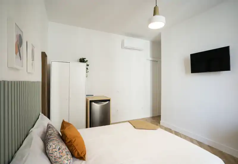 Dormitorio luminoso y minimalista con cama grande vestida en blanco, cojines de color y televisión en pared — líneas limpias y luz suave que crean un ambiente tranquilo para descansar.