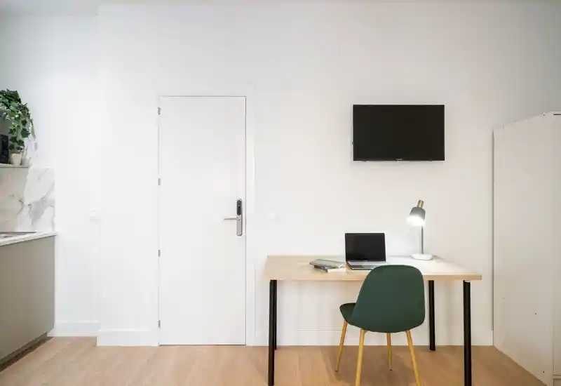 Zona de estar minimalista con escritorio, silla y televisor de pared; paredes blancas y suelo de madera que ofrecen un espacio moderno y funcional, ideal para estancias cortas o estudios.