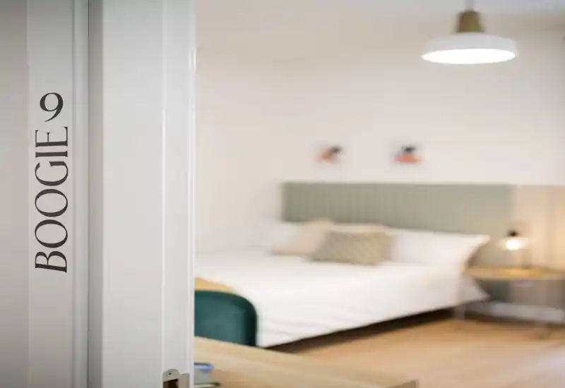 Dormitorio moderno y luminoso fotografiado a través de una puerta entreabierta, con cama doble bien arreglada, cabecero acolchado, mesita y luz ambiental suave.