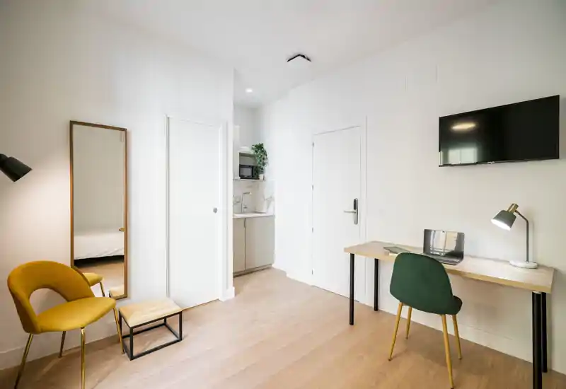 Estudio/cama luminoso y moderno con escritorio, silla y cama visible por una puerta abierta — suelos de madera y líneas limpias que crean un espacio acogedor.