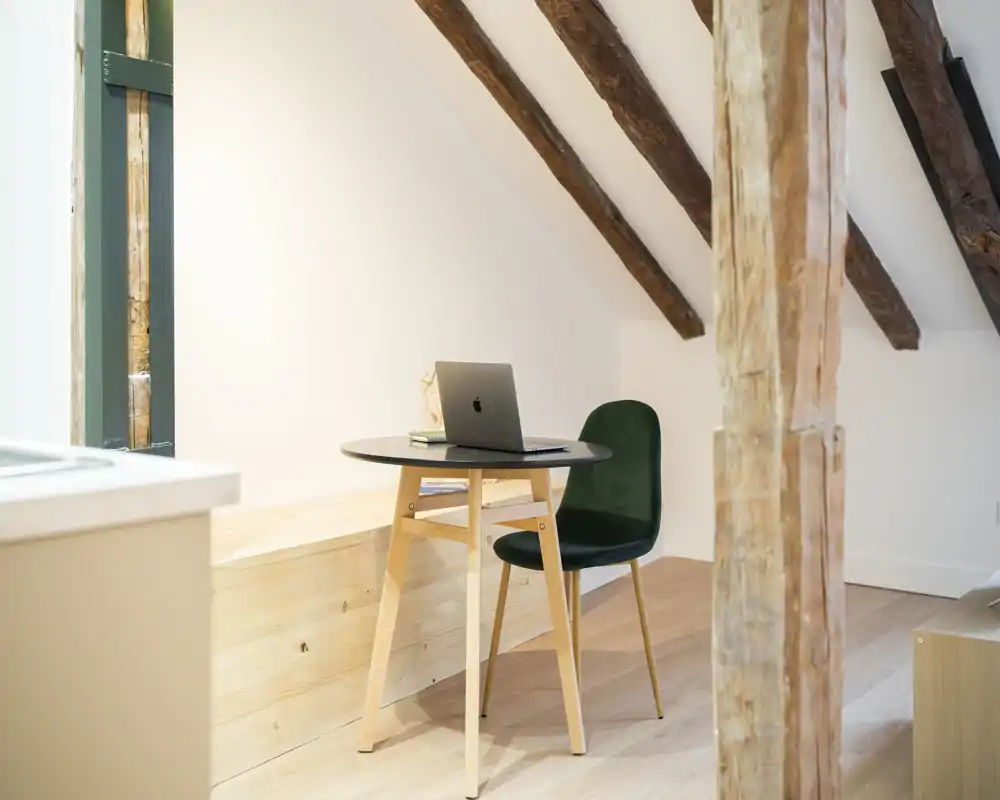 Angolo luminoso e minimal sotto travi in legno a vista, con un piccolo tavolo rotondo e una sedia verde imbottita — perfetto come zona soggiorno compatta o area lavoro.