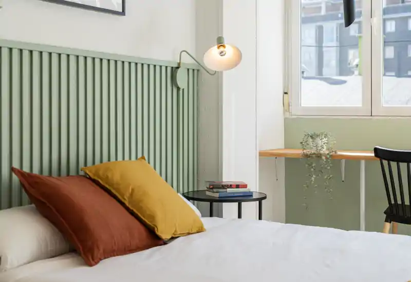 Bright, simply styled bedroom with a bed dressed in pillows and soft textiles by a window; small bedside table and light contribute to a cozy, inviting feel.