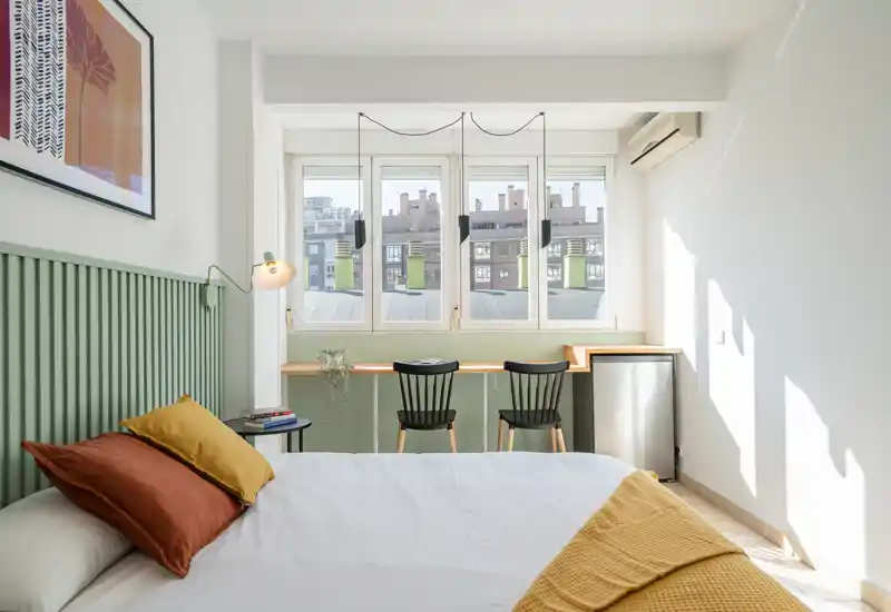 Bright, styled bedroom with a clearly visible bed, decorative pillows and a cozy throw, set against a patterned headboard and a window letting in natural light.