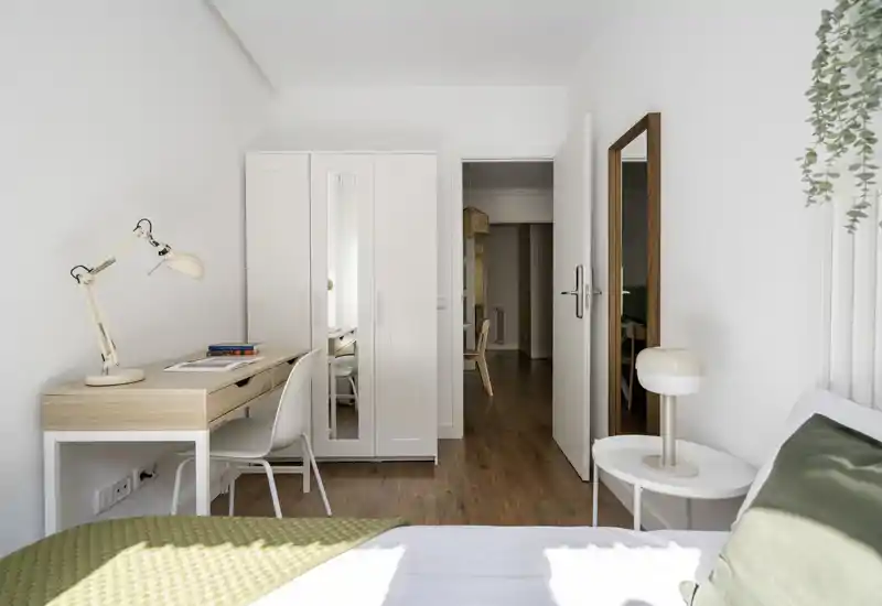 Camera da letto luminosa e minimal con un comodo letto, comodino e una scrivania in legno chiaro vicino alla finestra; stile scandinavo neutro e luce naturale che creano un'atmosfera tranquilla.