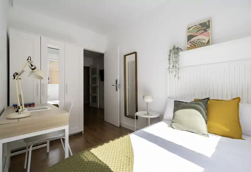 Camera da letto luminosa e ordinata con testiera bianca, cuscini decorativi senape e oliva, comodino e scrivania accanto all'armadio. La luce naturale illumina l'ambiente, creando un'atmosfera fresca e accogliente perfetta per annunci di affitto.