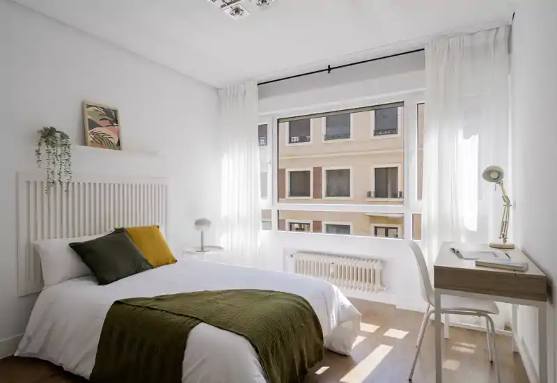 Camera da letto luminosa e minimal con letto completo, tessili morbidi e una grande finestra che lascia entrare molta luce naturale.