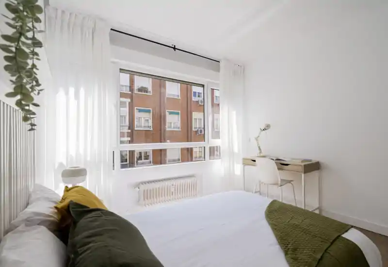 Camera da letto luminosa e minimale con una grande finestra, biancheria bianca e una scrivania compatta: ambiente ideale per riposo o lavoro da remoto.