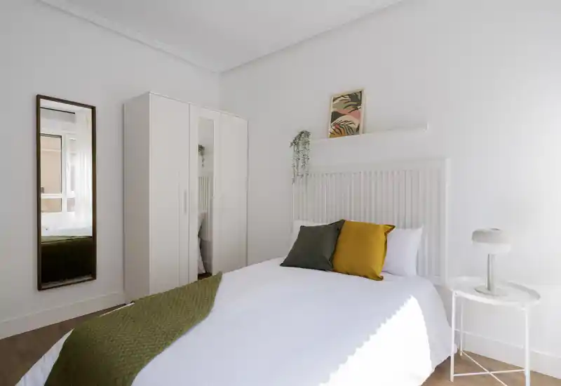 Camera da letto minimalista e luminosa con letto ben rifatto, cuscini verdi e ocra, un armadio e uno specchio a figura intera; pareti bianche e pavimento in legno donano un'atmosfera tranquilla.