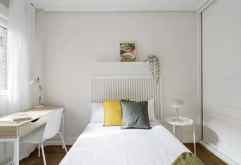 Camera da letto luminosa e minimal con letto singolo centrale, cuscini decorativi, un piccolo comodino e una scrivania vicino alla finestra — pulita, ariosa e curata.