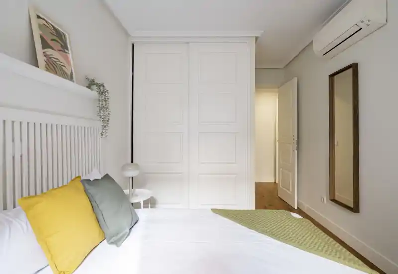 Camera da letto luminosa e ben arredata con testiera bianca, cuscini colorati e letto ben rifatto; dotata di armadio a muro e specchio a figura intera, perfetta per annunci.