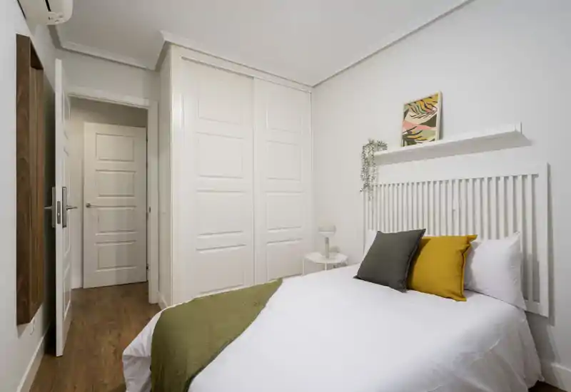 Camera da letto luminosa e ordinata con testiera a listelli bianca, cuscini giallo senape e grigio e un plaid verde sul piumone bianco. Armadi a muro e pavimento in legno chiaro completano l’atmosfera moderna e rilassante.