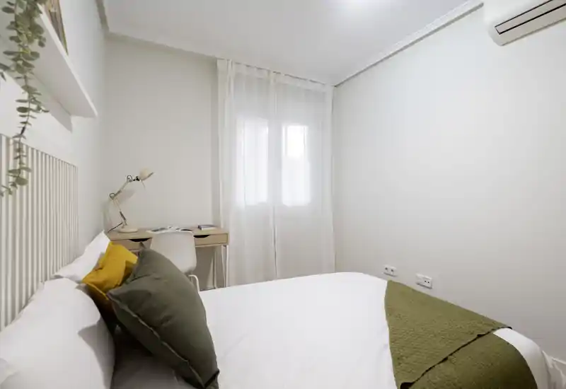 Camera da letto luminosa e minimale con letto matrimoniale ben rifatto, cuscini decorativi e una piccola scrivania vicino alla finestra — ideale per riposo e lavoro leggero.