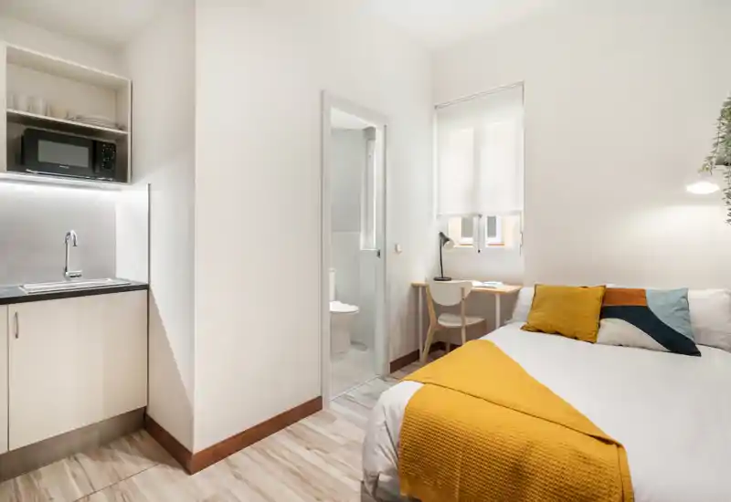 Bright, compact bedroom with a comfortable double bed, bedside lamp and a small desk by the window; kitchenette visible to the left and an en-suite bathroom door in the center.