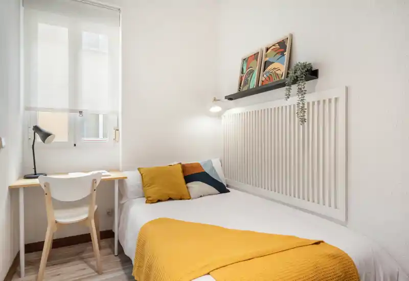 Compact, bright bedroom with a single bed dressed in white linens and mustard accents, a small workspace by the window, and simple modern decor for a cozy stay.