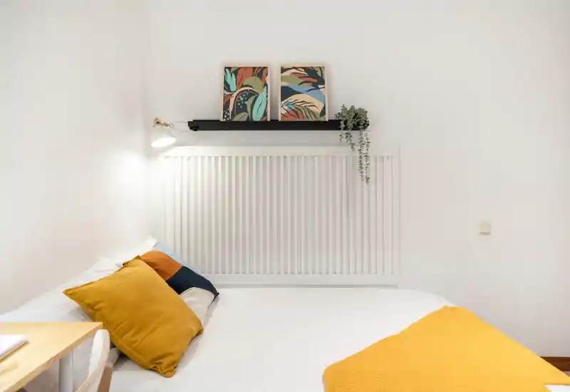 Bright, minimal bedroom with a large white bed, colorful cushions and a warm mustard throw; framed prints and a small plant add a modern, cozy touch.