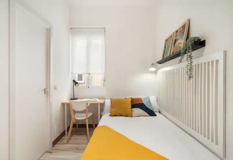 Cozy single bedroom with a comfortable bed against a white slatted headboard, decorative cushions and a yellow throw. A small desk and chair sit by the window, ideal for working, with a bedside wall lamp and shelf with plant accents.