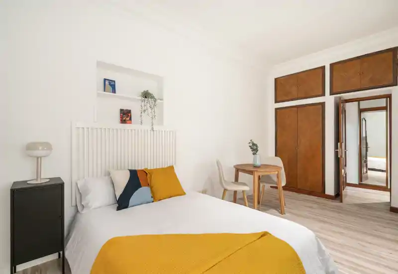 Bright, styled bedroom with a neatly made double bed, decorative cushions and a warm mustard throw. Wooden built-in wardrobes and a small table create a cozy, well-lit space ideal for short-term rentals.