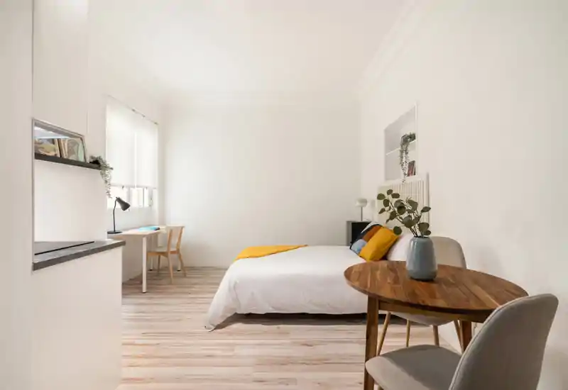 Bright, minimalist bedroom with a double bed, warm accent textiles and a small dining table near the bed. Large window provides abundant natural light; clean wooden floors and neutral walls create a calm, modern atmosphere.