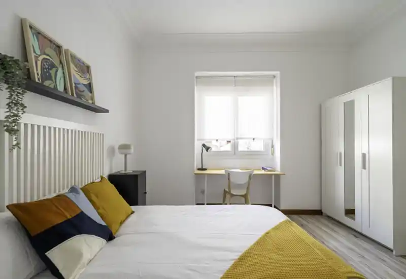 Bright, modern bedroom with a large white bed dressed in colorful cushions and a mustard throw, a desk by the window and a spacious wardrobe — airy, stylish and well lit.