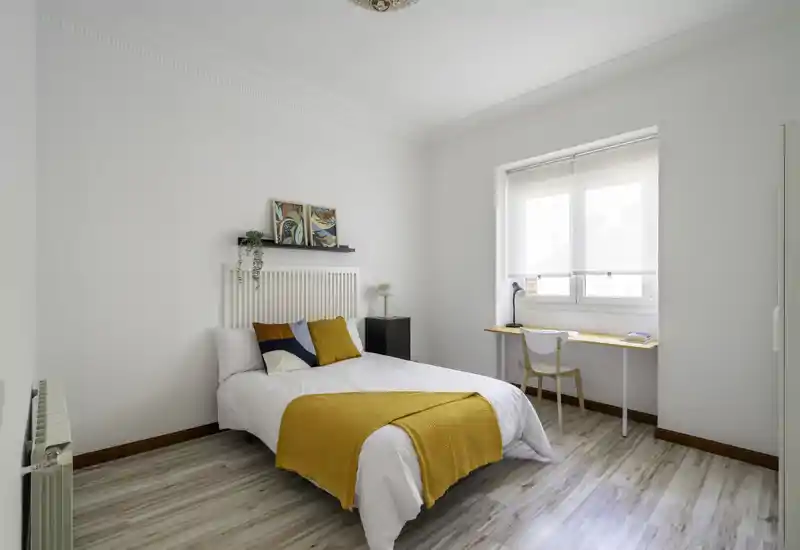 Bright, minimalist bedroom with a comfortable double bed dressed in white linens and a mustard throw, a small bedside unit and a window-side desk that receives abundant natural light.
