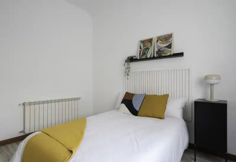 Bright, stylish bedroom with a neatly made double bed, colorful cushions and a mustard throw. Minimalist white walls, small bedside cabinet and tasteful wall art create a calm, modern atmosphere.