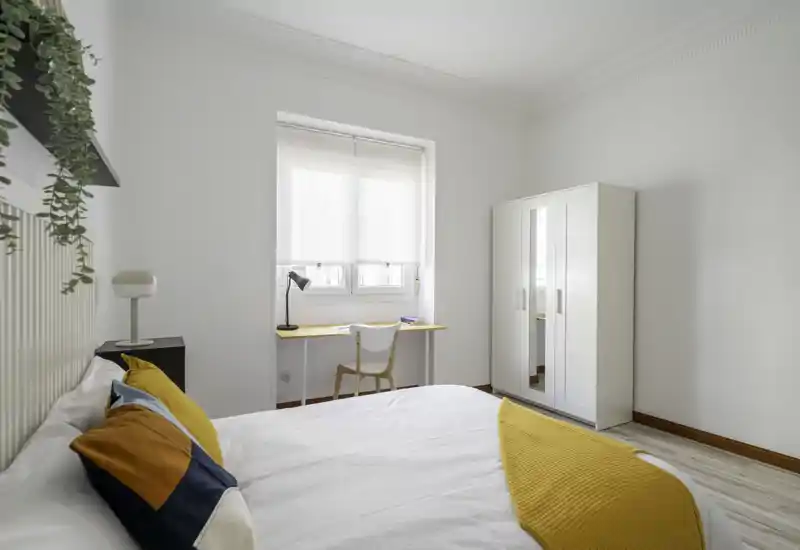 Chambre lumineuse et minimaliste avec un lit bien fait, des coussins colorés et un plaid jaune ; un petit bureau près de la fenêtre et une armoire complètent l'agencement fonctionnel.