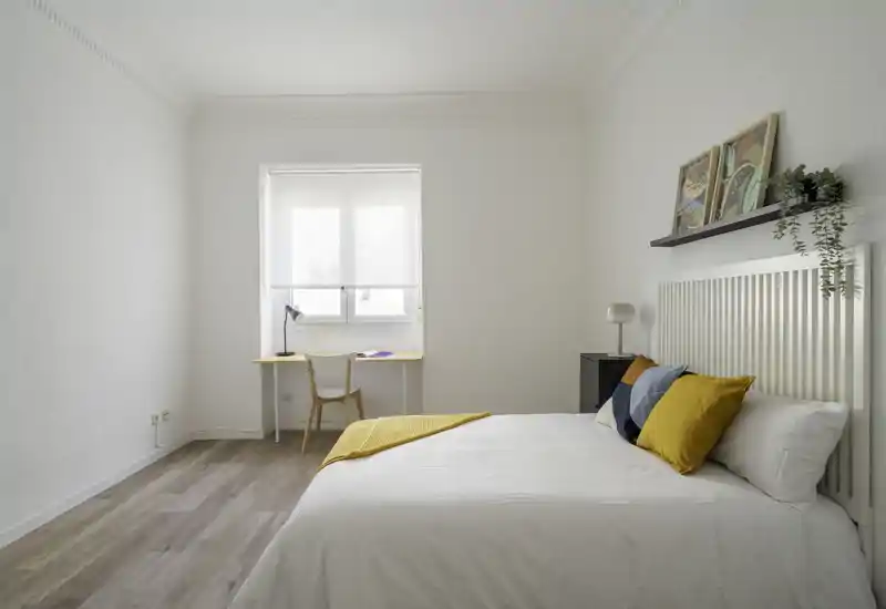 Bright, minimalist bedroom with a comfortable double bed dressed in white linens, colorful cushions and a yellow throw. A small desk sits under the window offering a natural-work corner; neutral walls and wood flooring create a calm, modern atmosphere.