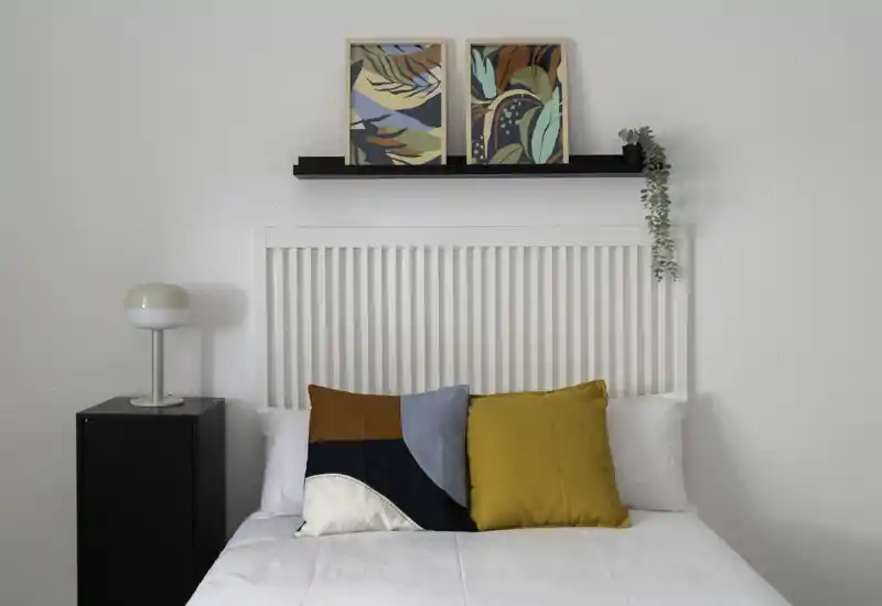 Bright, styled bedroom with a white slatted headboard, decorative cushions and framed art above the bed — clean, modern and welcoming.
