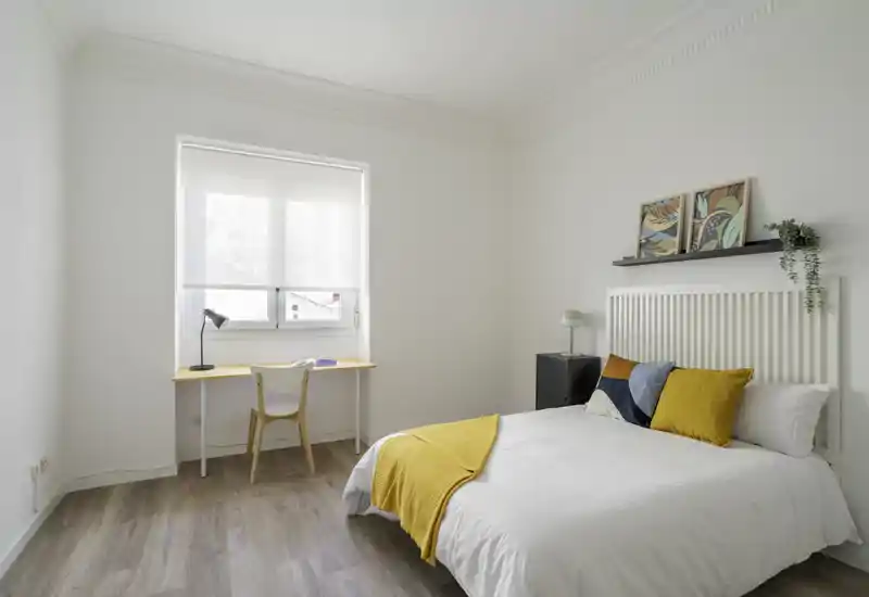 Bright, minimal bedroom with a comfortable double bed dressed in white linens and yellow accents, a simple desk by the window and natural light creating an airy atmosphere.